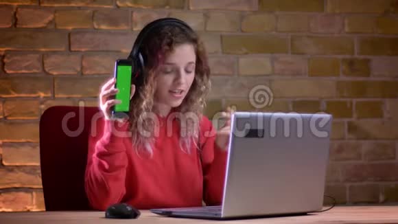 穿红色连帽衫的年轻女性博客作者的肖像展示了智能手机在briken墙背景下的笔记本电脑视频的预览图