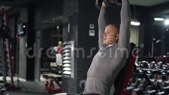 近距离强壮的肌肉男正在黑暗的健身房训练运动员坐在长凳上按哑铃训练视频的预览图