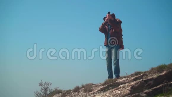 戴牛仔帽皮夹克蓝色牛仔裤肩上背着旅游背包的人一个人透过望远镜看视频的预览图