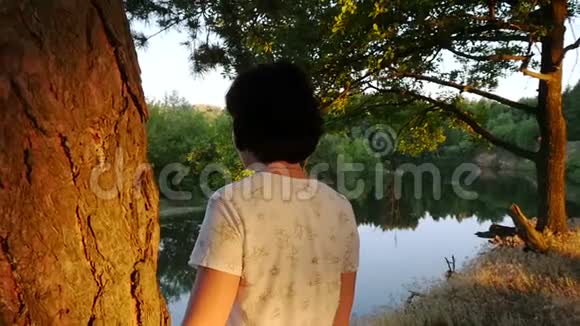 一个年轻的女人站在森林和河流背景上的一棵树附近作为相机的圆周运动视频的预览图