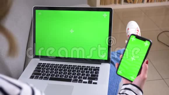 特写后景拍摄女性使用笔记本电脑和手机绿色屏幕坐在舒适的室内沙发上视频的预览图