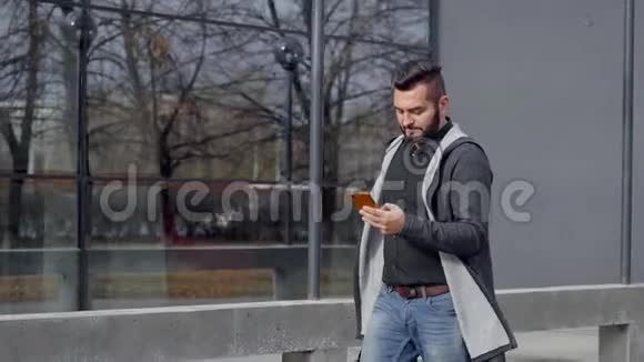 迷人的胡须男人走着微笑着看着他的智能手机背着一个袋子视频的预览图