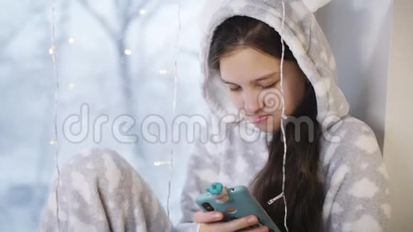 女孩少年冬天穿着温暖的睡衣坐在窗边用你的智能手机给朋友发短信窗外视频的预览图