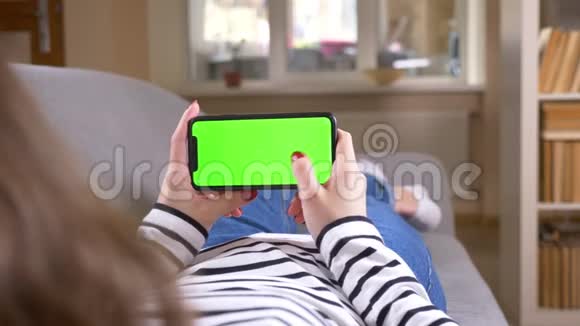 特写后景拍摄女性滚动手机与绿色屏幕保持它水平躺在沙发上视频的预览图