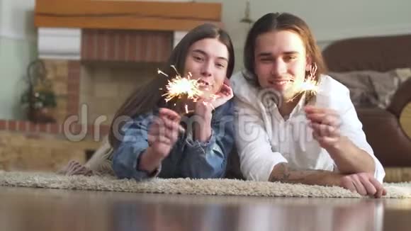 年轻漂亮的快乐男孩和女孩手里拿着火花躺在家里的地毯上视频的预览图
