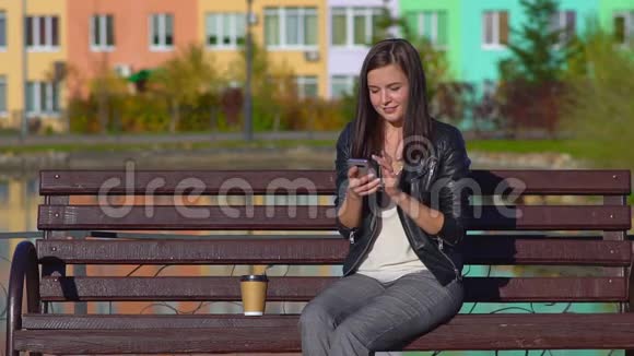 公园里一个年轻迷人的女孩坐在长凳上收到短信看书和笑视频的预览图