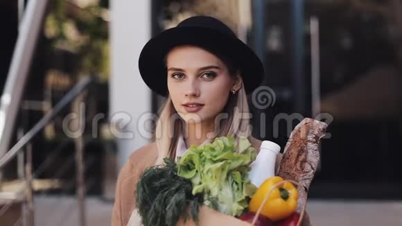 年轻漂亮的女人穿着时髦的外套站在城市街道上手里拿着一包微笑和微笑的产品视频的预览图