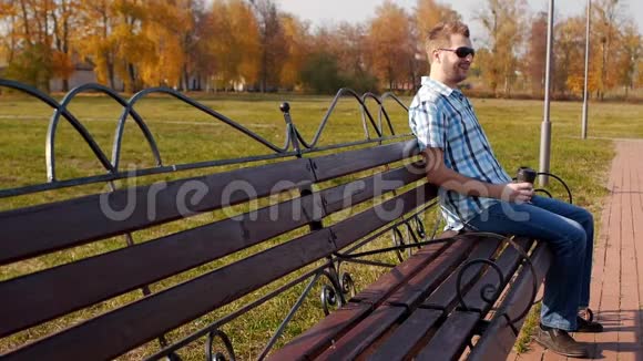 一个戴着黑眼镜的年轻白种人坐在公园里的长凳上喝咖啡慢节奏时尚复制空间视频的预览图