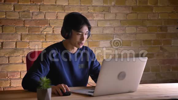 一位年轻的韩国商人戴着耳机在室内笔记本电脑上打视频电话的特写照片视频的预览图