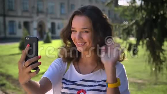 一位年轻微笑的白种人女孩站在公园里用智能手机打招呼视频聊天制作的肖像视频的预览图