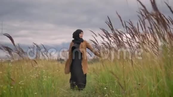 年轻美丽的穆斯林妇女戴着头巾用手触摸田野中的植物穿过草地视频的预览图