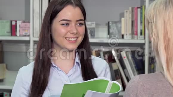 可爱的年轻女子笑着和她在大学图书馆的朋友说话视频的预览图