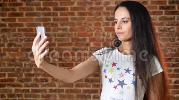 年轻可爱的女孩打招呼视频聊天智能手机微笑砖墙在背景肖像视频的预览图