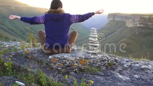 女孩坐在莲花的位置在山上的岩石上日落后景冥想和自然的概念视频的预览图