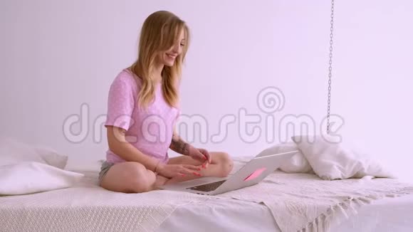 一个漂亮的金发女孩穿着一件欧洲外观的粉红色T恤拿着笔记本电脑坐在床上在家工作聊天聊天视频的预览图