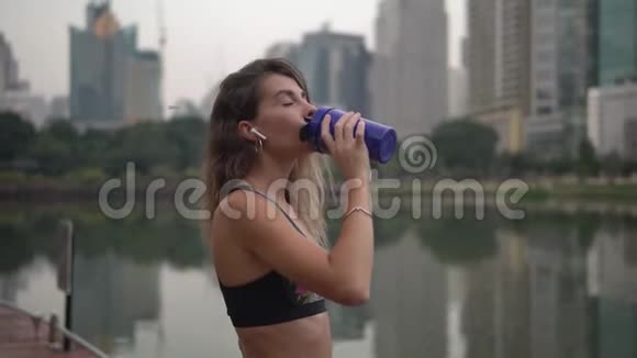 年轻的黑发女人在公园里跑步后在蓝色健身杯里喝健身饮料视频的预览图