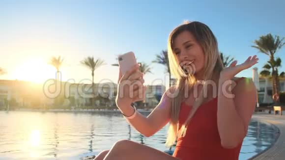 年轻女孩在豪华别墅的游泳池附近放松通过智能手机与朋友在线视频通话视频的预览图