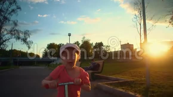 小可爱的金发女孩穿着粉红色的帽子在公共公园骑滑板车视频的预览图
