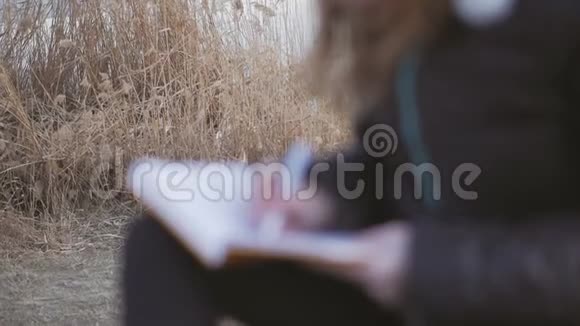 春天的下午一个穿着深色夹克的年轻女孩坐在河边用笔在笔记本上记笔记女人视频的预览图