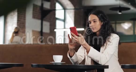 漂亮的年轻商务女性穿着粉红色西装使用智能手机在舒适的咖啡馆或现代办公室喝咖啡视频的预览图