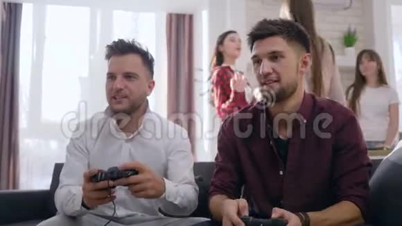 年轻的游戏玩家朋友男性坐在沙发上玩电子游戏而有趣的女友则是派对的背景视频的预览图