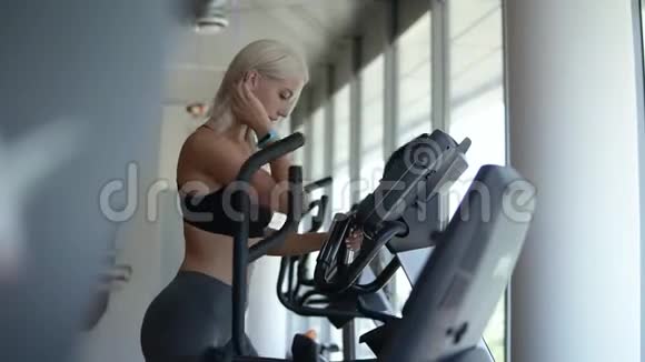 一个穿着运动服的金发女孩在明亮的空健身房的椭圆教练锻炼的侧面有全景窗慢慢视频的预览图