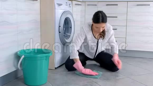 戴着粉色橡胶手套的疲惫女人用一块布洗厨房地板看着摄像机地板上的灰色瓷砖视频的预览图