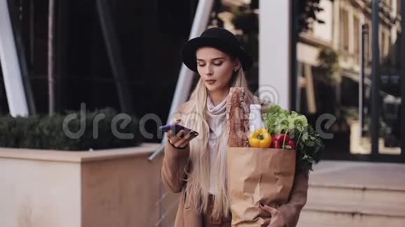 穿着时髦外套的年轻漂亮女人站在街上拿着一包产品和说话视频的预览图