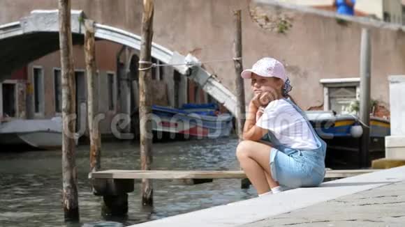 意大利威尼斯2018年7月7日悲伤的女孩穿着短裤和帽子坐在一条小运河的岸边靠近大桥视频的预览图