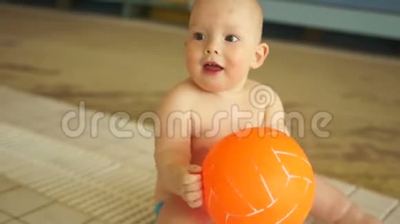 可爱的宝宝正在玩橙色的球坐在温暖的孩子们水上乐园的游泳池里视频的预览图