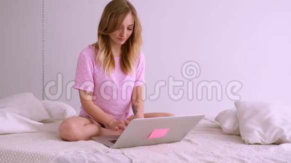 一个漂亮的金发女孩穿着一件欧洲外观的粉红色T恤坐在床上拿着笔记本电脑在家工作聊天聊天视频的预览图