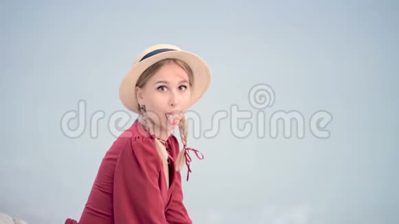 一位美丽的白种人年轻女子身穿红色连衣裙头戴草帽头戴辫子坐在坐在海滨微笑视频的预览图