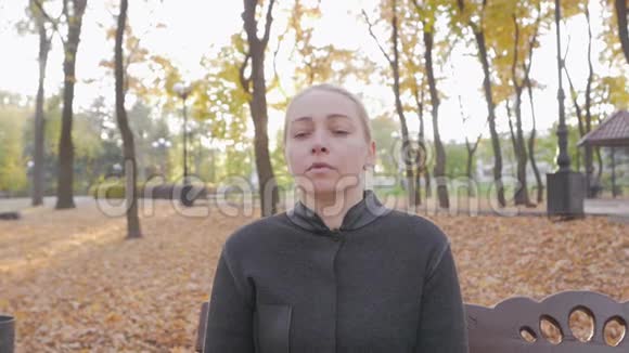 一个女人坐在一个城市秋天公园的长凳上手里拿着她的头头痛视频的预览图