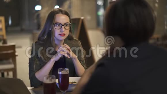 戴着眼镜的漂亮年轻女人和她的朋友在户外咖啡馆聊天视频的预览图