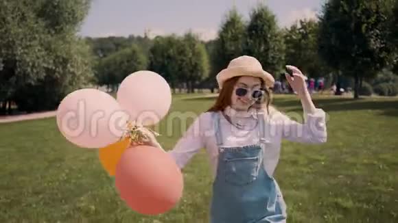 戴着帽子的开朗的姜女在公园里用气球跑步摄像视频的预览图