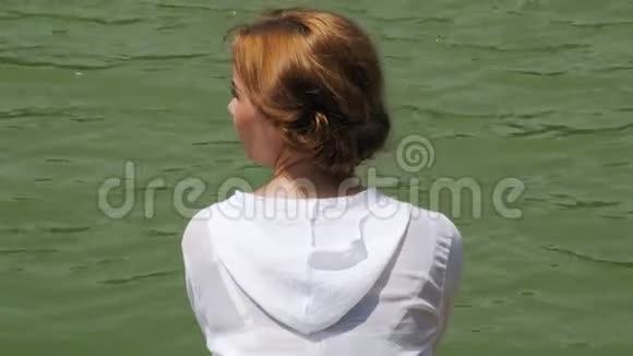 穿着白衬衫的女孩在江水背景上享受夏日年轻女子在绿湖岸边放松看着视频的预览图