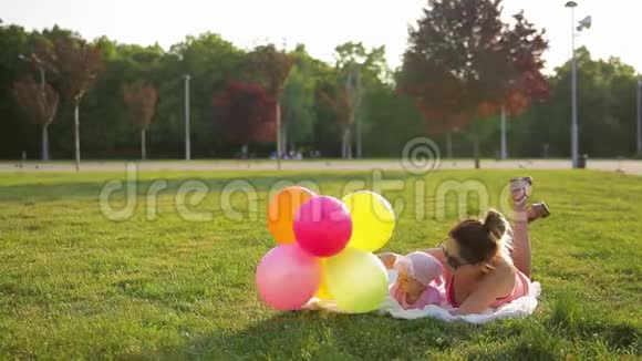 年轻的母亲和可爱的小女儿穿着粉红色的衣服躺在铺在绿色草地上的白色毯子上视频的预览图