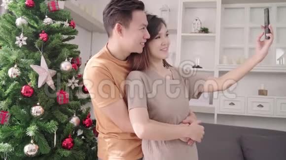亚洲夫妇使用智能手机自拍与圣诞树装饰他们的客厅在家里圣诞节视频的预览图