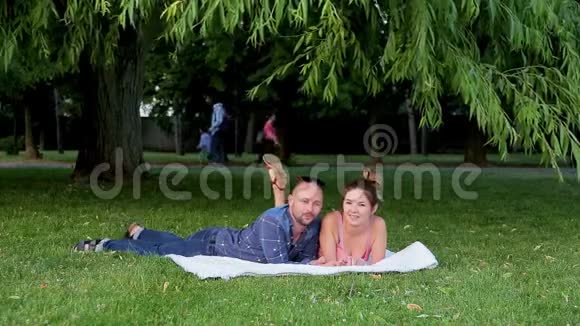 一对漂亮的夫妇躺在铺在绿草上的白色毯子上在茂密的树叶下闲聊着视频的预览图