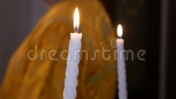 新娘和新郎手中拿着两支燃烧着的白色蜡烛在一位牧师的东正教背景下的草地上燃烧视频的预览图