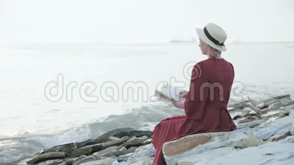 日落时分一个漂亮的白人女孩穿着红色鲜艳的裙子和一顶草帽坐在海边的一块大石头上视频的预览图