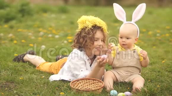 两个兄弟一个少年和一个婴儿正在一个空地上的公园里休息复活节概念兔子耳朵复活节篮子和鸡蛋视频的预览图