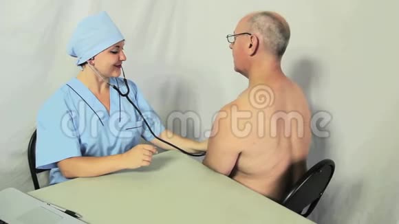一位女医生坐在他的办公桌前用听诊器倾听男人胸部镜头移向小车视频的预览图