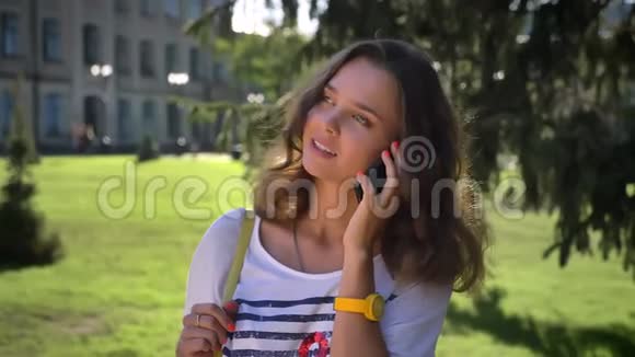 一位年轻微笑的白种人女孩站在公园里在智能手机上说话滚动着大学里视频的预览图