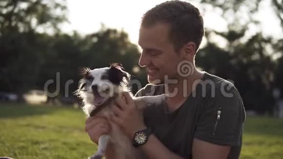 男人坐在绿色公园的草地上和年轻的小狗玩主人逗狗狗玩和拥抱微笑快乐视频的预览图