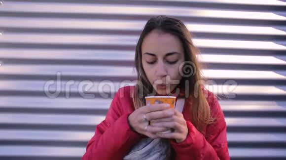 年轻女子穿着红色夹克穿着蓝色牛仔裤腰袋在户外的金属墙背景上喝咖啡视频的预览图
