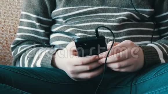 一个少年男孩的手他拿着黑色智能手机坐在沙发上听耳机里的音乐视频的预览图