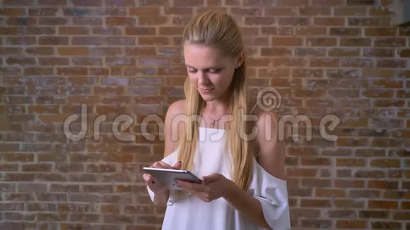 年轻可爱的白人金发女人用平板电脑刷卡看着相机微笑着背景是砖墙视频的预览图