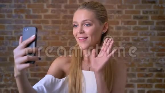 年轻可爱的白种人金发女孩在视频聊天中打招呼挥动她的手打招呼的手势砖墙在里面视频的预览图