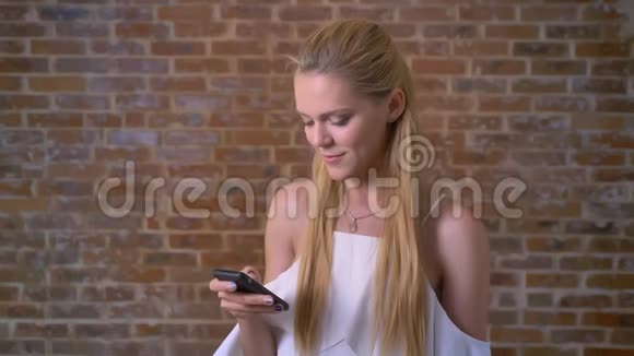 年轻可爱的白种人金发女郎使用智能手机微笑着背景是砖墙视频的预览图
